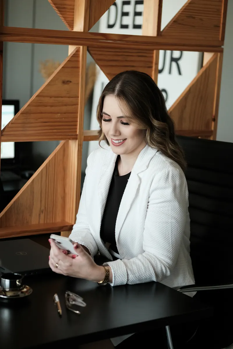 Estér Coelho sorrindo à mesa de trabalho, segurando o celular